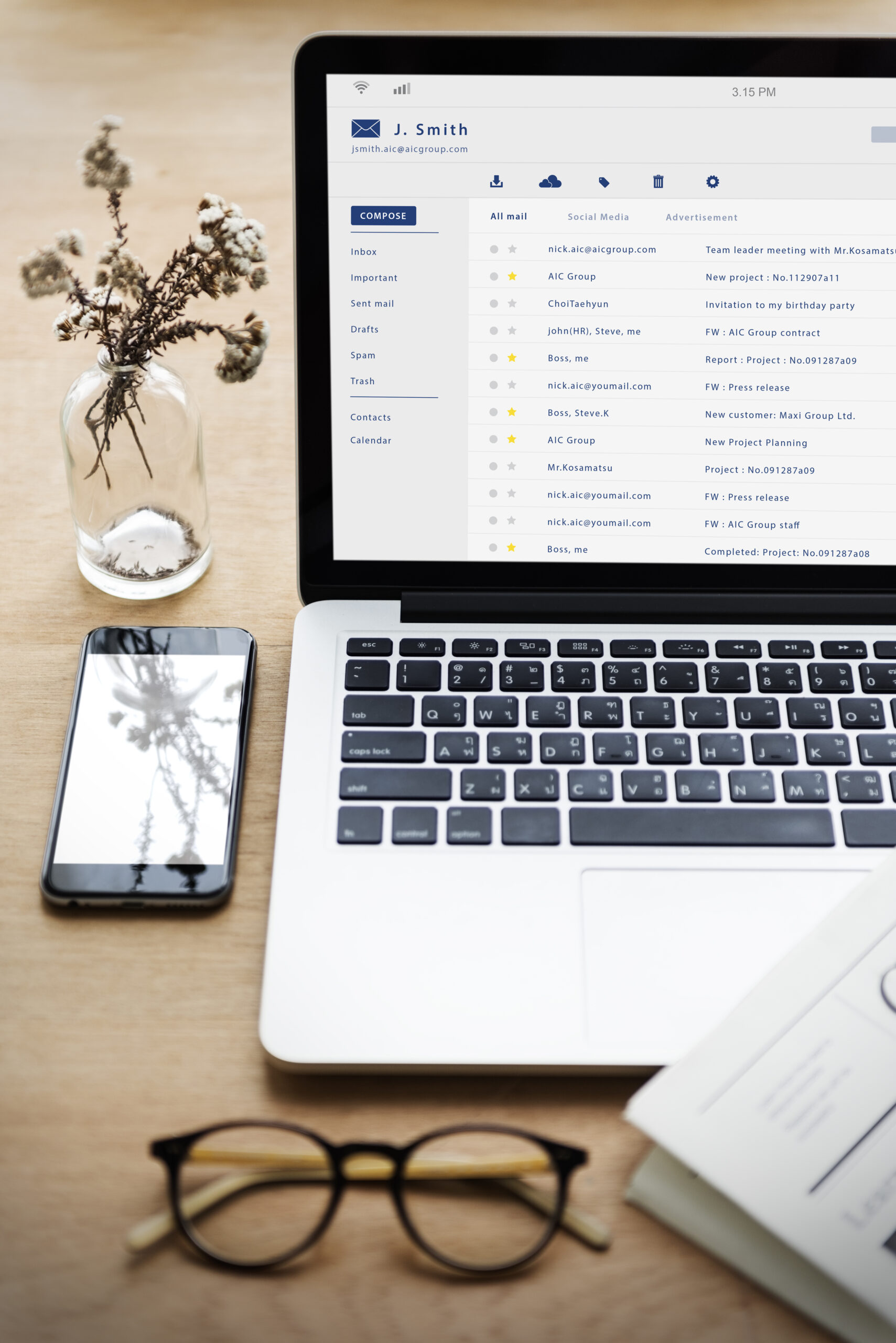 a laptop open to email, sitting on a desk with a phone, glasses and a small vase